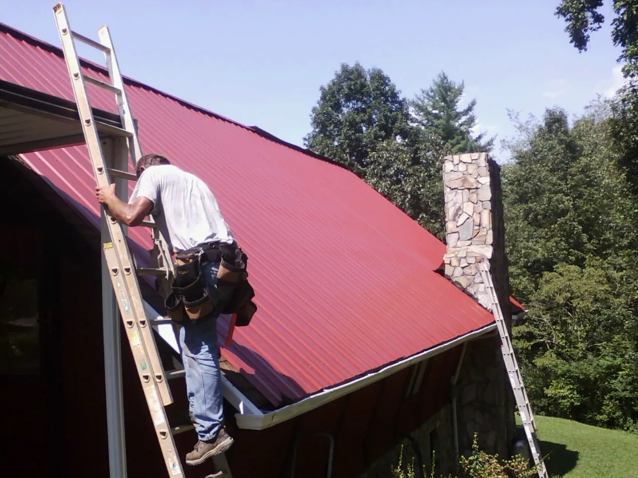 Roofer on a ladder beside a red metal roof and stone chimney — Soffit & Fascia Repair by Roofing Brothers in Arkadelphia
